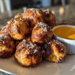 A pile of golden brown soft pretzel bites sprinkled with coarse salt, served next to a ramekin of cheese sauce.
