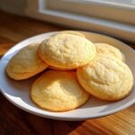 Close-up of several soft sugar cookies, slightly puffed and coated in granulated sugar, stacked on a white plate.