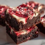 Two stacked, fudgy candy cane brownies topped with a white and red marbled cream cheese swirl and crushed peppermint pieces.