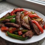 Close-up of sizzling steak fajitas featuring strips of seasoned beef, red and green bell peppers, and onions on a white plate.