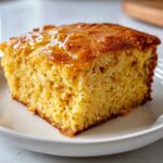 Close-up of a square slice of moist sweet potato cornbread topped with a shiny glaze, served on a white plate.