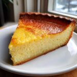 A close-up slice of golden brown buttermilk pie with a slightly sunken, glossy top, served on a white plate.