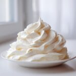 A tall, perfectly piped swirl of creamy white vanilla buttercream frosting on a small white plate.