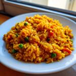 A close-up of vibrant, yellow Spanish rice mixed with green peas and diced red vegetables served on a white plate.