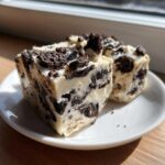 Two thick squares of white chocolate oreo fudge loaded with crushed dark chocolate cookies, served on a small white plate.