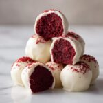 A stack of white chocolate coated red velvet cake balls, with several cut open to show the deep red interior.