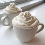 Two white mugs filled with creamy white hot chocolate, topped generously with swirls of whipped cream.
