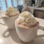 Two white mugs filled with rich white hot chocolate topped generously with swirls of whipped cream.