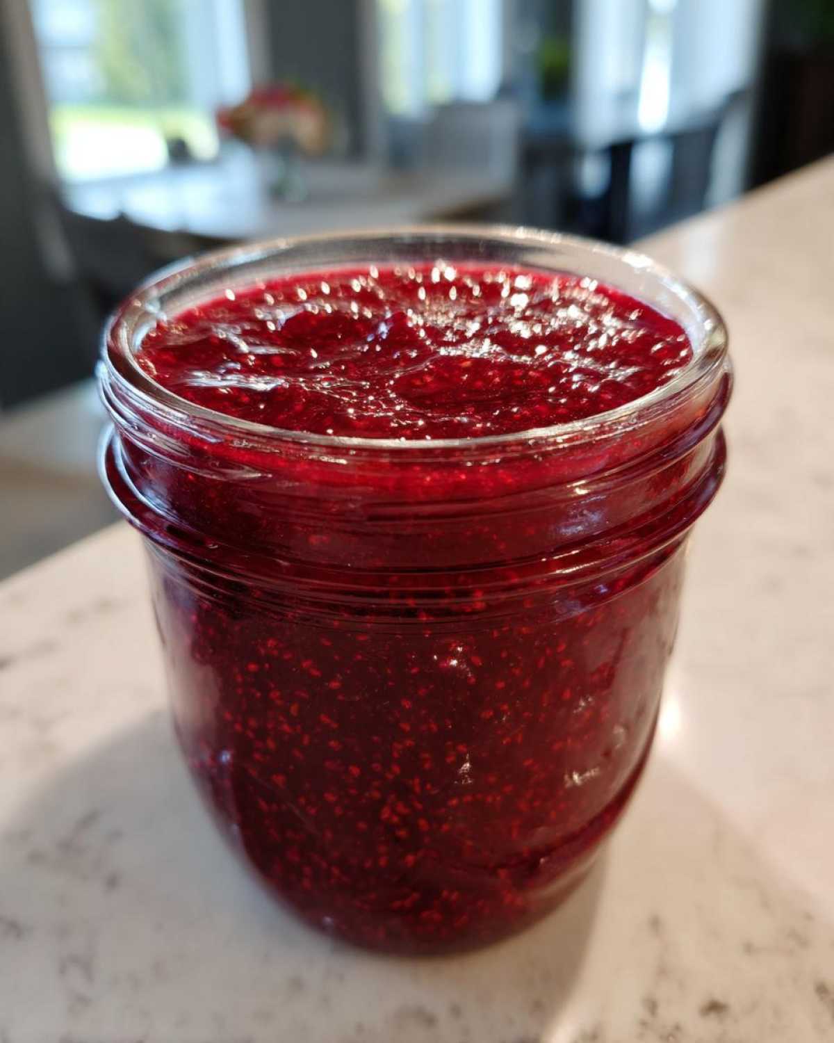 A close-up of a glass jar filled with vibrant, thick homemade raspberry jam, showing visible seeds.