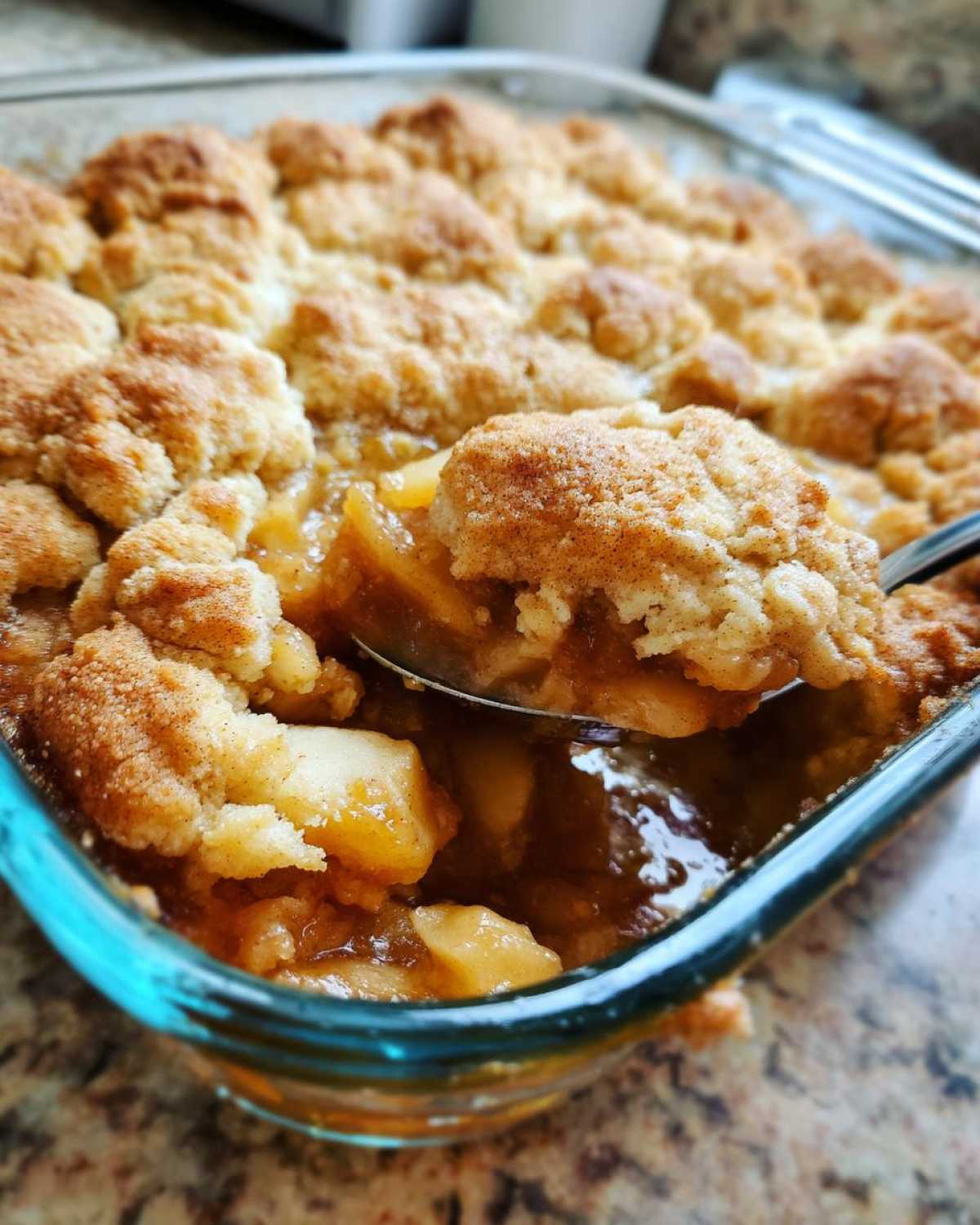 Close-up of a spoonful being lifted from a glass dish of amazing apple cobbler recipe, showing warm, spiced apples and crumb topping.