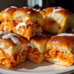 Close-up of several gooey buffalo chicken sliders piled on a white plate, topped with a white sauce drizzle.