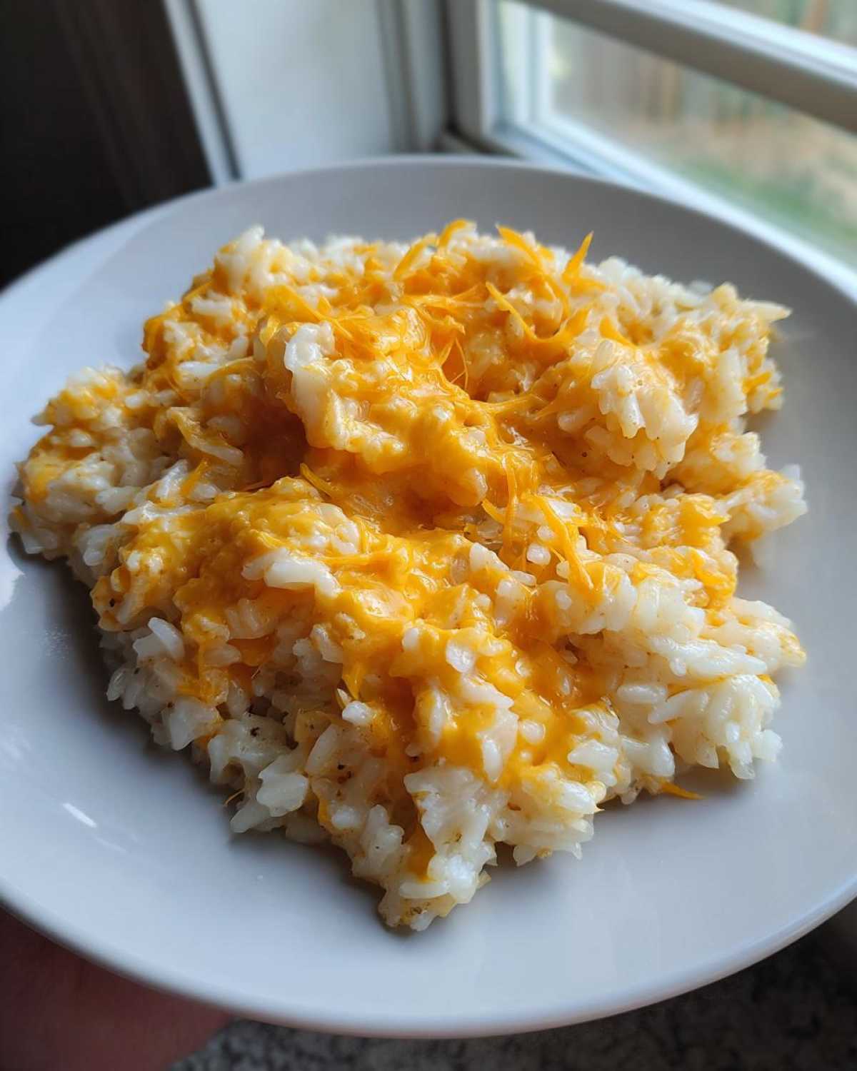 A close-up of a serving of amazing cheesy rice topped with melted cheddar cheese on a white plate.