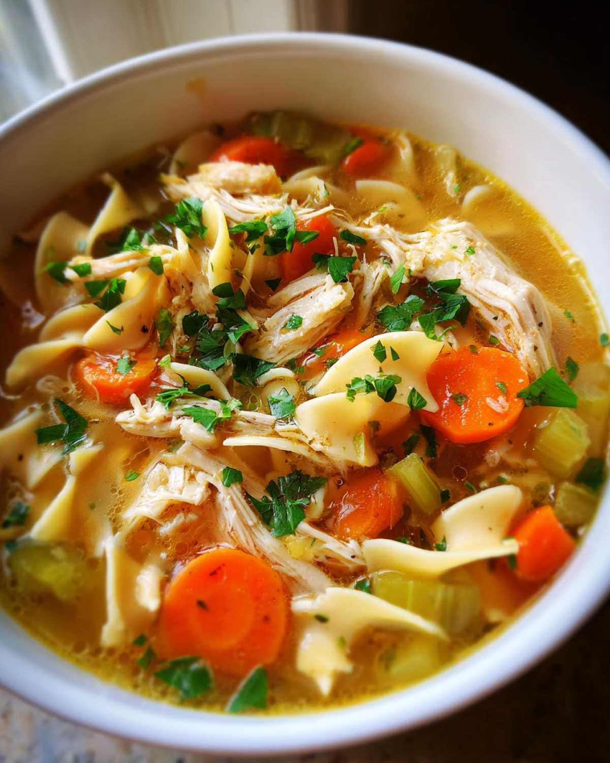 Close-up of a bowl of amazing chicken noodle soup, featuring shredded chicken, egg noodles, sliced carrots, and celery, garnished with parsley.