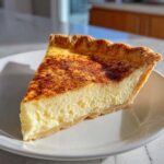 A close-up of a perfect slice of creamy custard pie dusted with cinnamon on a white plate.