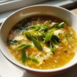 A close-up of a white bowl filled with golden yellow egg drop soup, topped generously with bright green sliced scallions.