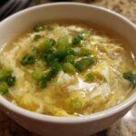 A close-up of a white bowl filled with hot egg drop soup, garnished generously with chopped green onions.