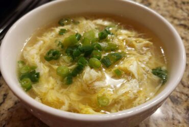 A close-up of a white bowl filled with hot egg drop soup, garnished generously with chopped green onions.