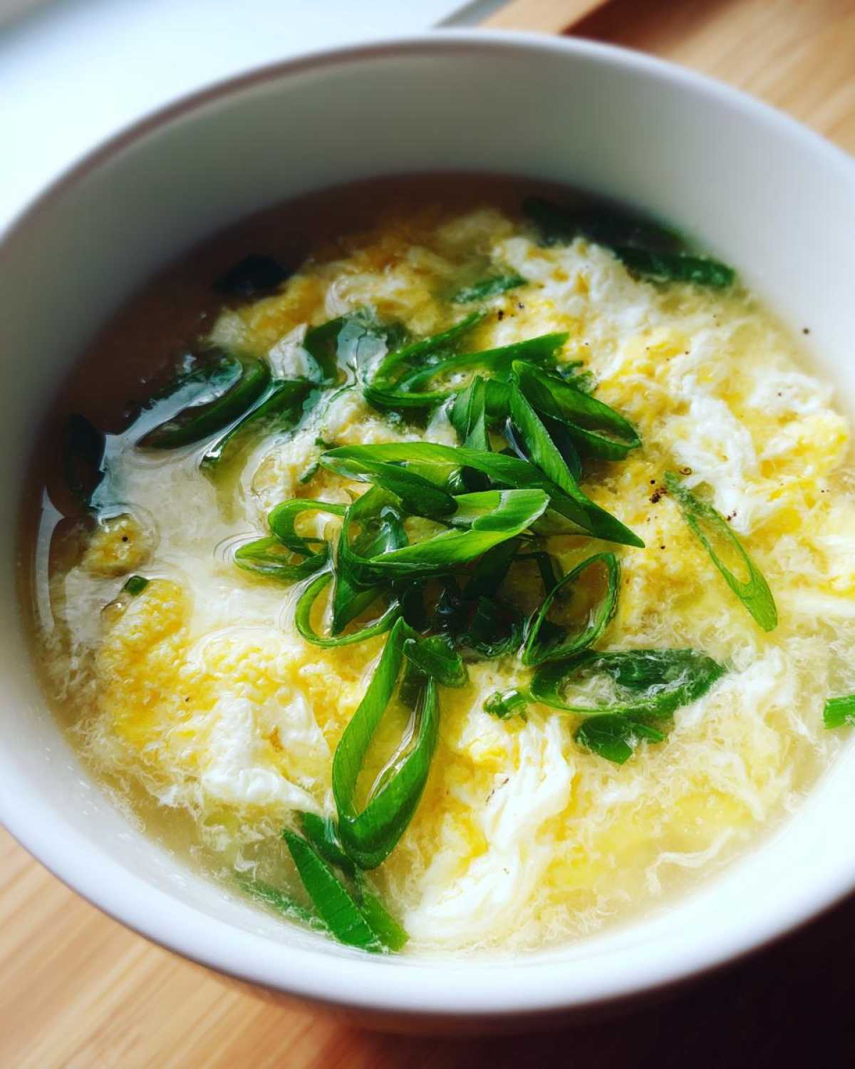 A close-up view of a white bowl filled with steaming egg drop soup, featuring wispy cooked egg ribbons and green onion garnish.