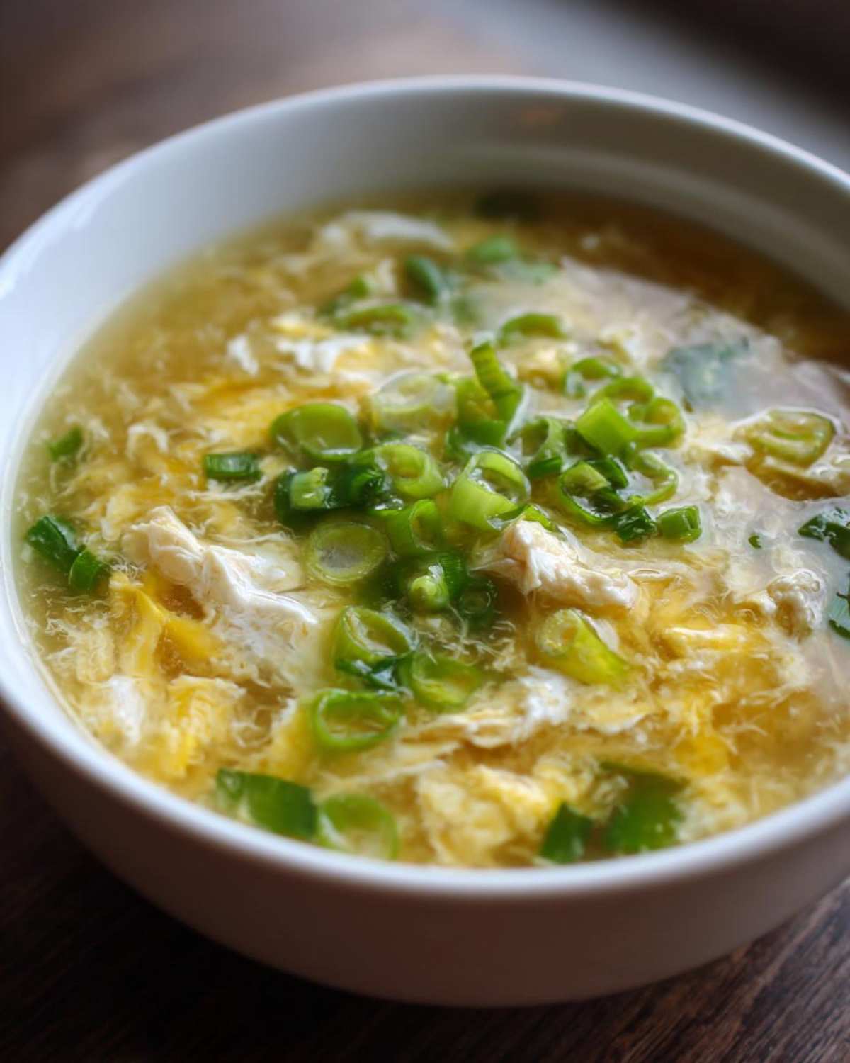A close-up of a white bowl filled with hot egg drop soup, featuring wisps of cooked egg and topped with fresh green onions.