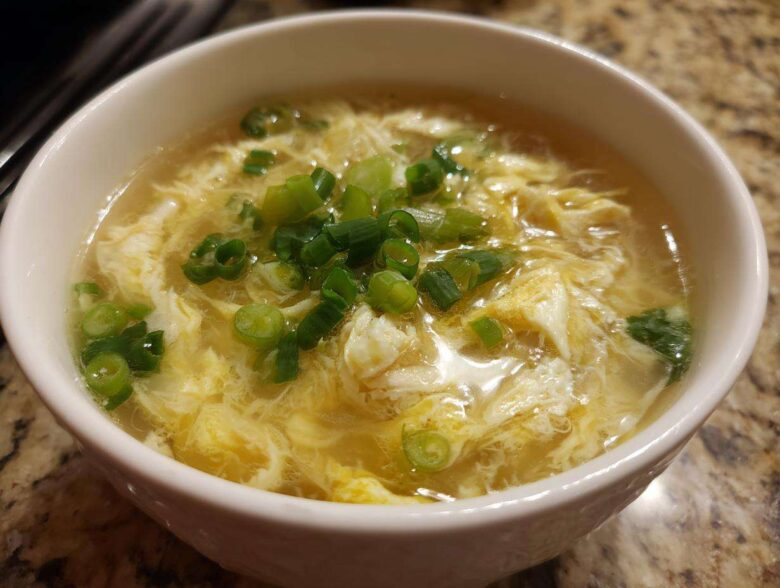 A close-up of a white bowl filled with hot egg drop soup, garnished generously with chopped green onions.