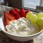 A white bowl filled with creamy fruit dip topped with fresh strawberries and green grapes.