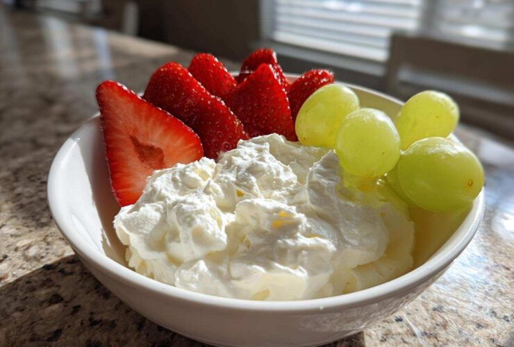 A white bowl filled with creamy fruit dip topped with fresh strawberries and green grapes.