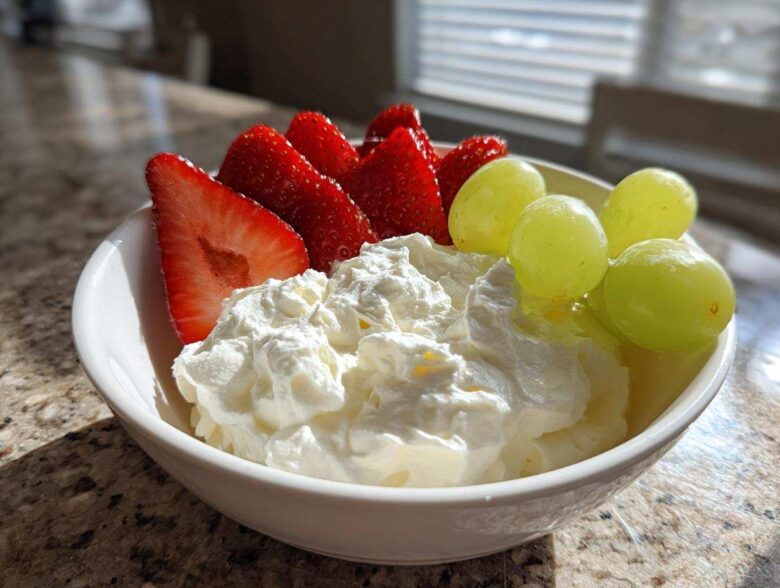 A white bowl filled with creamy fruit dip topped with fresh strawberries and green grapes.