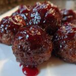 A close-up of several savory Grape jelly meatballs coated in a thick, shiny, dark red glaze on a white plate.