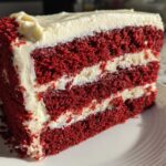 A tall, moist slice of red velvet cake featuring three layers of deep red cake separated by thick layers of white cream cheese frosting.