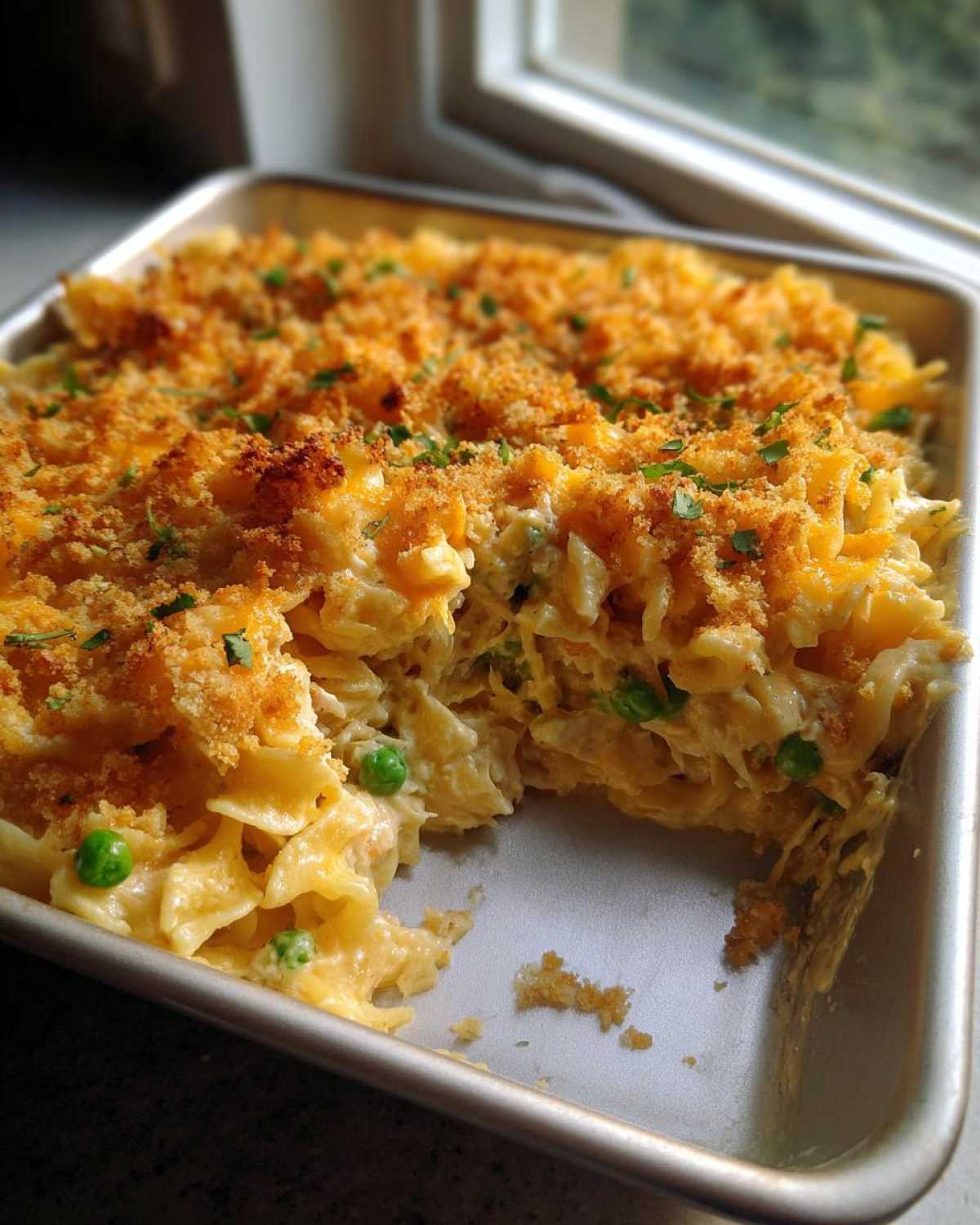 A creamy, baked Tuna Casserole with egg noodles and peas, topped with golden breadcrumbs, served in a metal pan.
