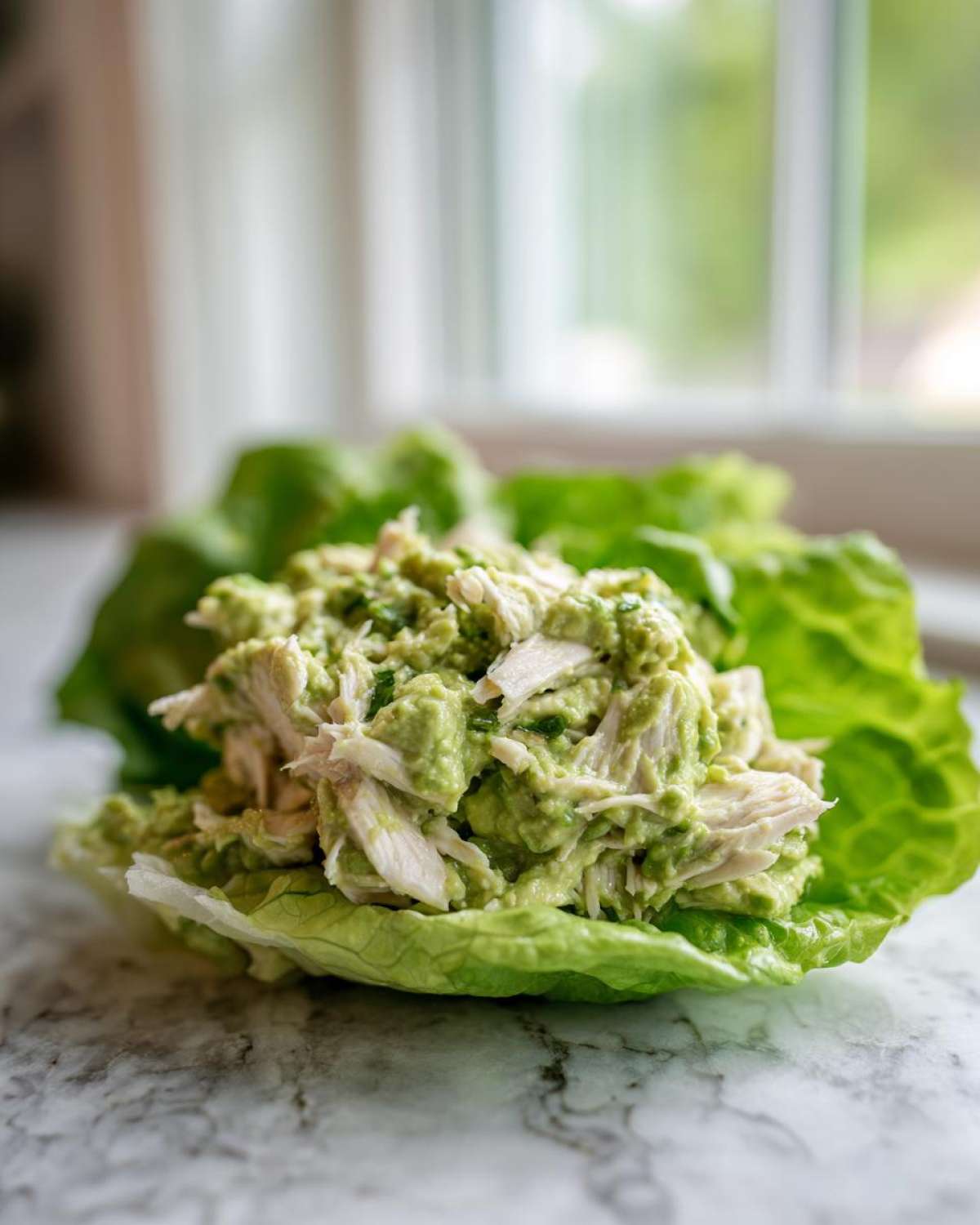 Close-up of creamy avocado chicken salad served in a crisp butter lettuce cup on a marble surface.