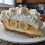 A generous slice of banana cream pie recipe featuring a graham cracker crust, creamy filling, fresh banana slices, and whipped cream.