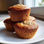 A stack of three golden-brown banana protein muffins resting on a white plate near a window.