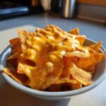 A close-up of tortilla chips generously covered in bright, creamy nacho cheese sauce, served in a small bowl.