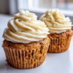 Two delicious carrot cake muffins topped with thick swirls of cream cheese frosting, sitting on a white surface.