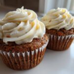 Close-up of two moist carrot cake muffins topped with thick swirls of cream cheese frosting.