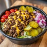 Close-up of a delicious cheeseburger bowl featuring seasoned ground beef, melted cheese sauce, pickles, tomatoes, and red onion over lettuce.