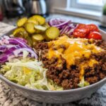 A close-up of a delicious cheeseburger bowl featuring seasoned ground beef topped with melted cheddar cheese, lettuce, red onion, pickles, and tomatoes.