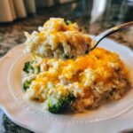 A spoonful of creamy cheesy chicken and rice mixed with broccoli being lifted from a serving on a white plate.