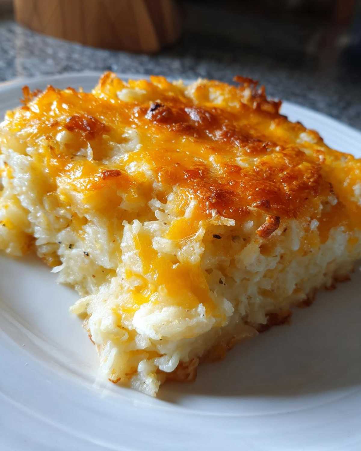 A close-up of a square serving of cheesy potato casserole topped with bubbly, golden-brown melted cheddar cheese.