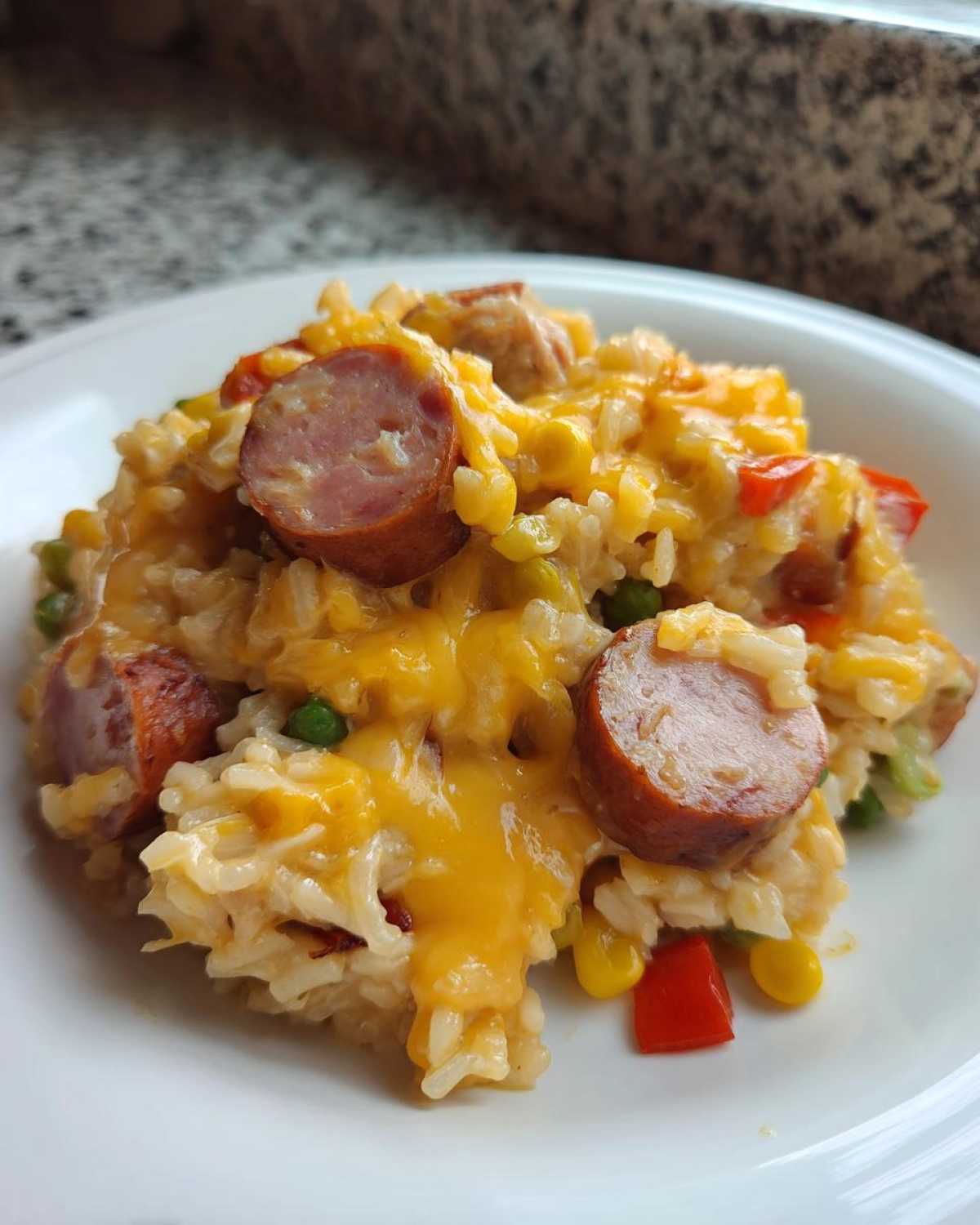 Close-up of a serving of cheesy sausage and rice mixed with corn, peas, and red peppers on a white plate.
