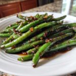 Close-up of bright green beans coated in a savory, dark chinese green beans with garlic sauce on a white plate.