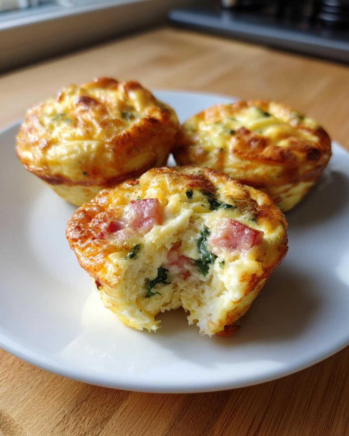 Three baked cottage cheese egg cups starbucks style on a white plate, one has a bite taken out showing ham and spinach.