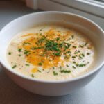 A white bowl filled with creamy cauliflower soup, topped with melted cheddar cheese and fresh chopped chives.