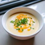 A close-up of creamy cauliflower soup in a white bowl, garnished with melted orange cheese and fresh chopped chives.