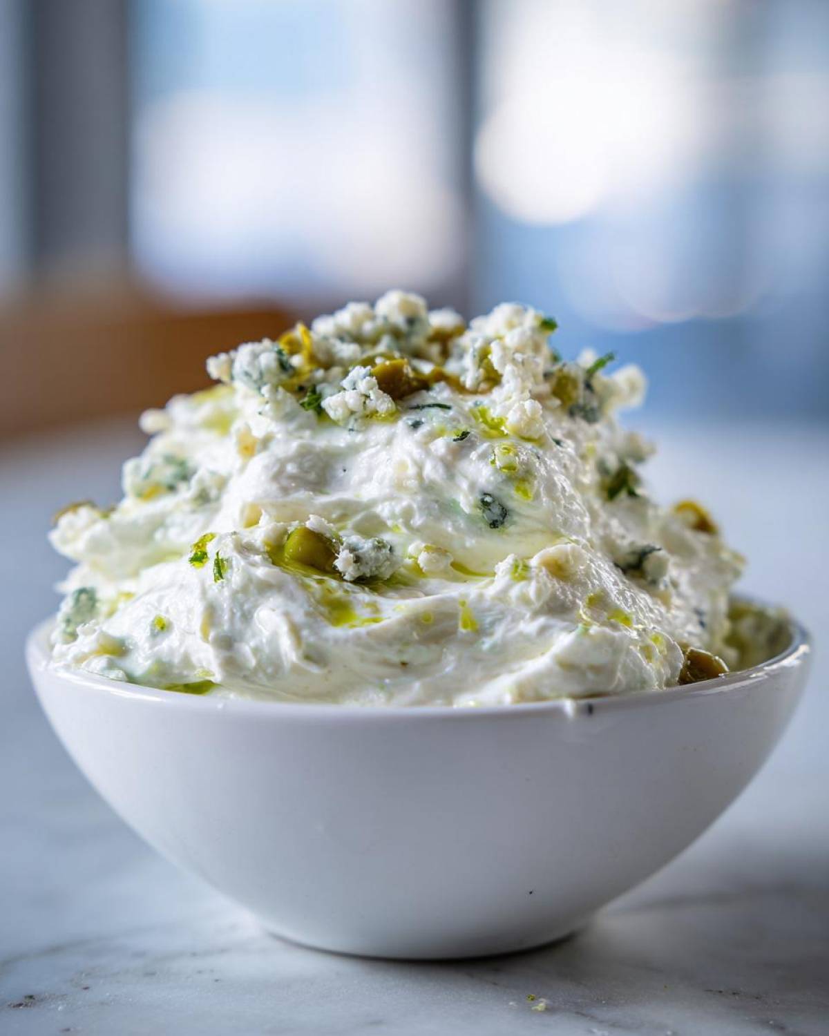 Close-up of a creamy, white dirty martini dip topped with blue cheese crumbles and chopped green olives.