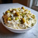 A bowl of creamy dirty martini dip topped with blue cheese crumbles, chopped celery, and whole green olives.