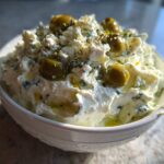 Close-up of a creamy, white dirty martini dip topped with crumbled cheese, green olives, and herbs.