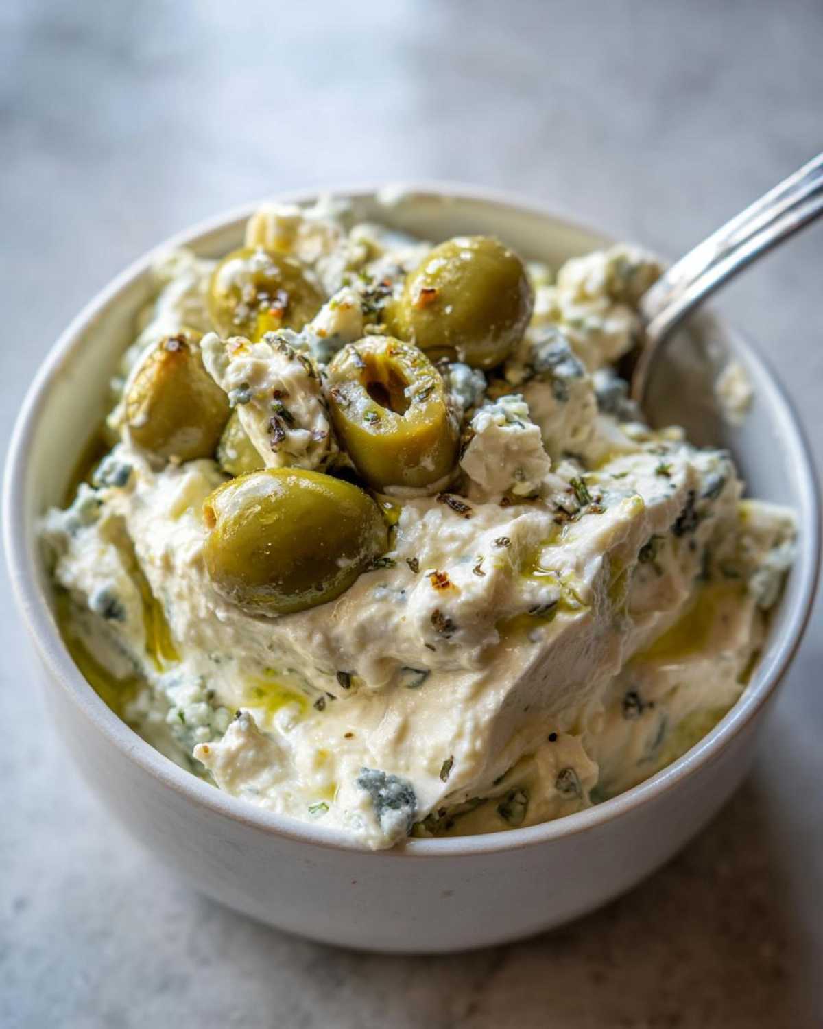 A close-up of the creamy dirty martini dip, topped with green olives, blue cheese crumbles, and a drizzle of oil.