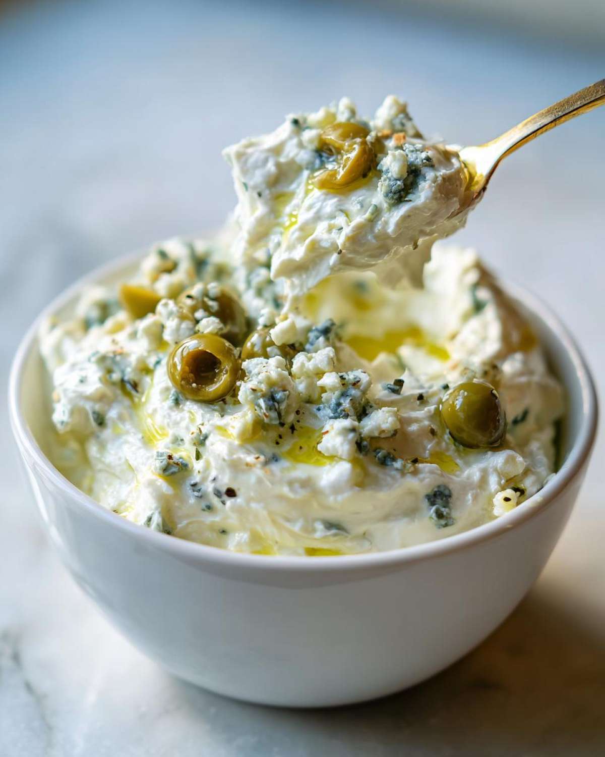 A golden spoon scoops a portion of creamy dirty martini dip topped with blue cheese crumbles and green olives.